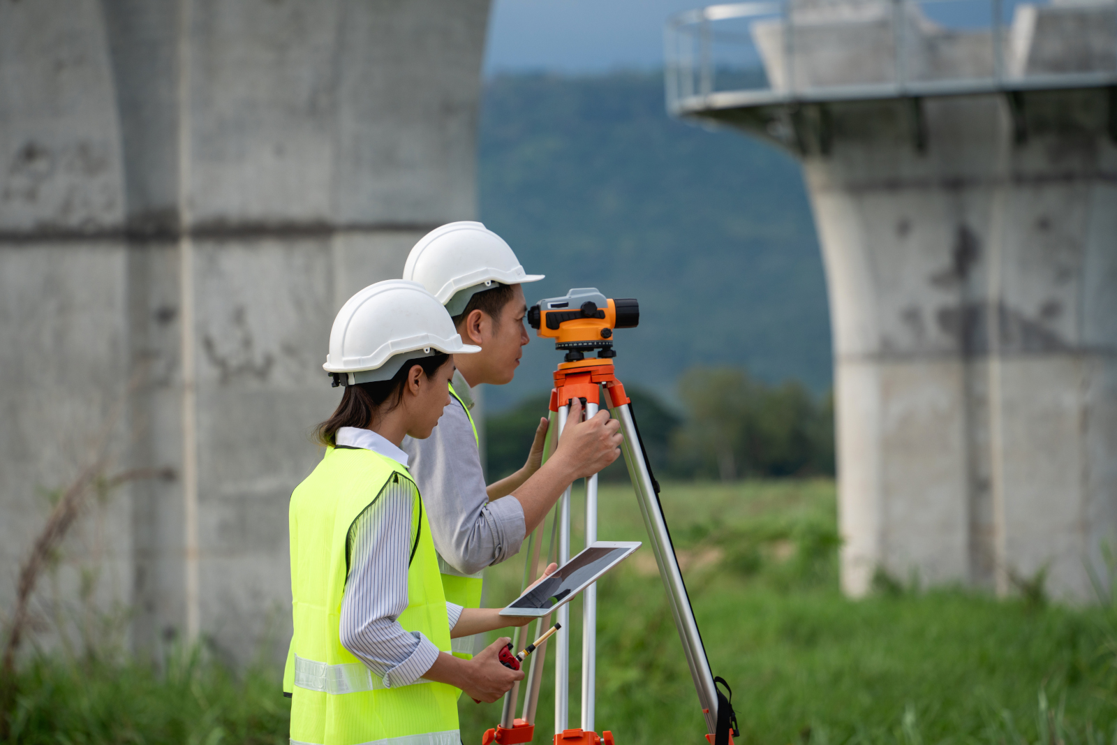 Male and female land surveyors in safety gear using theodolite and the NestForms Land survey app on smart device