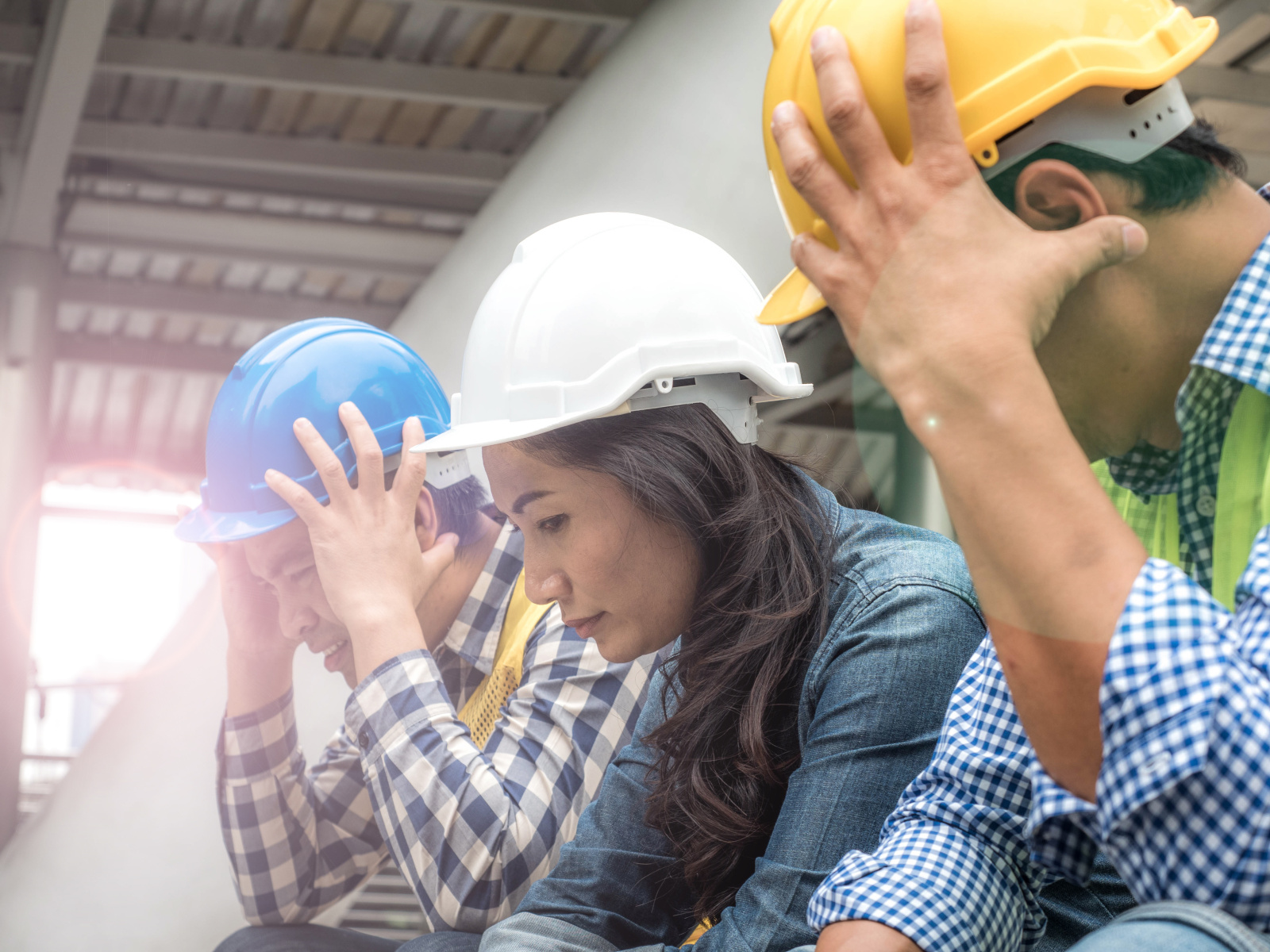 Construction Workers Looking Stressed on Urban Jobsite, three workers sat down and wearing hardhats holding their heads.