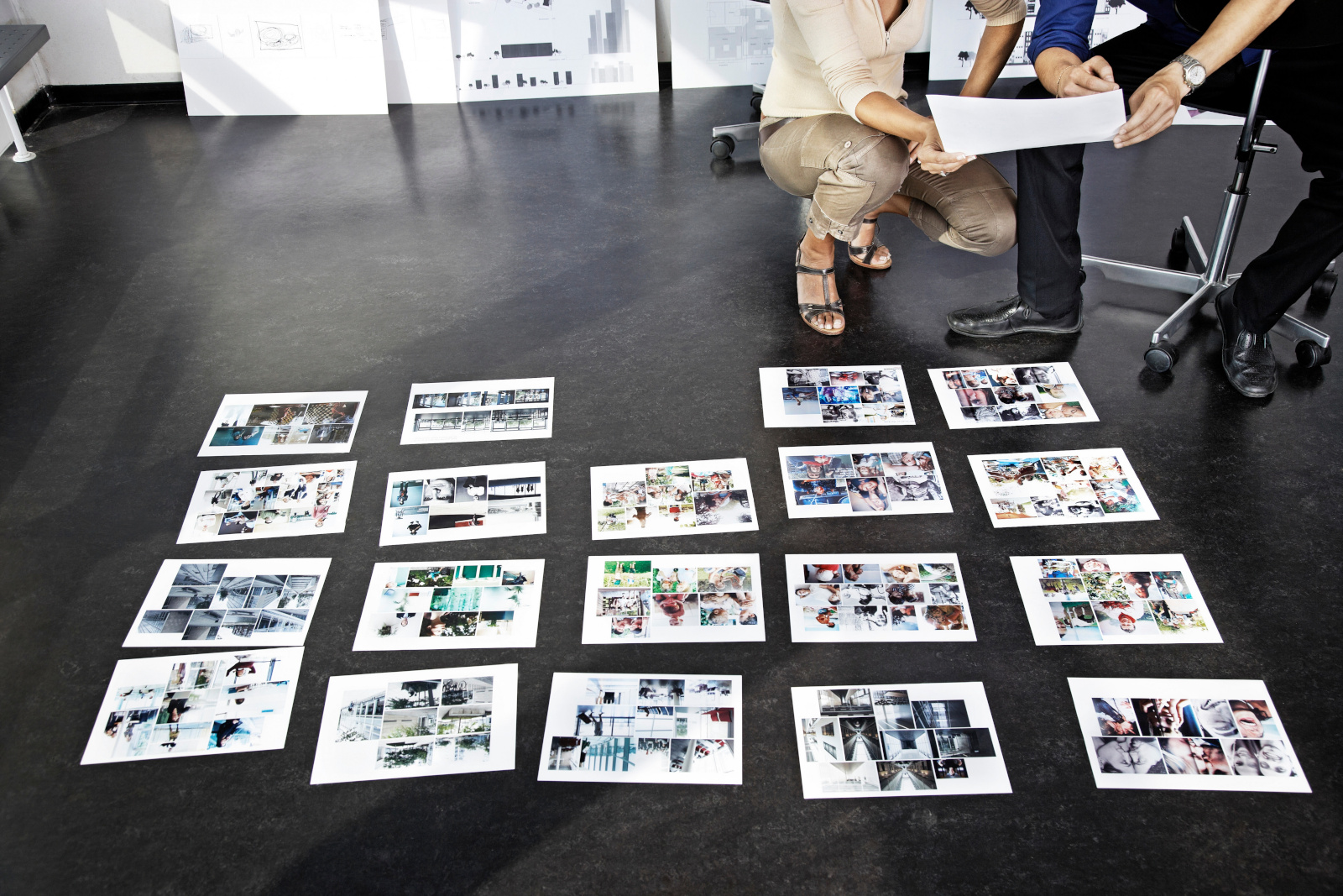 Two people are looking at a collection of photographs that are spread out on a dark floor.The photographs all depict different scenes. The light in the scene is bright and natural.  